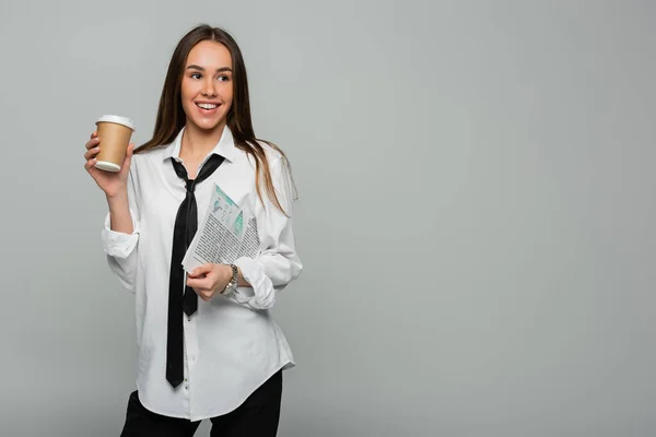 Cheerful woman in white shirt with tie holding newspaper and paper cup with coffee to go isolated on grey — Stock Photo
