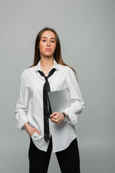 Young brunette woman in white shirt with tie holding laptop while standing with hand in pocket isolated on grey — Stock Photo