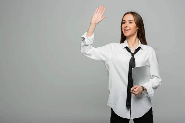 Happy woman in white shirt with tie holding laptop while waving hand isolated on grey — Stock Photo