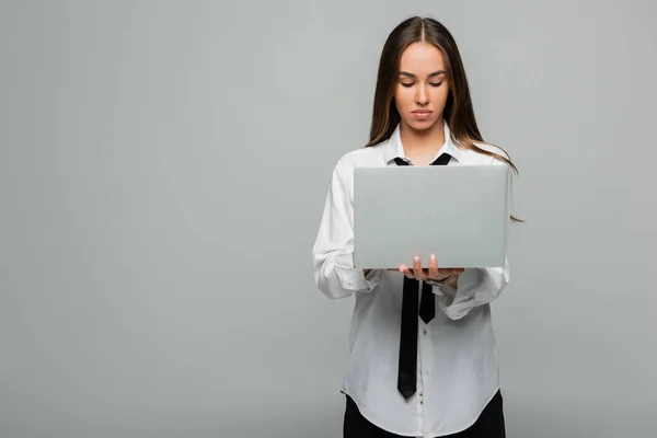 Young brunette woman in white shirt with tie using laptop isolated on grey — Stock Photo