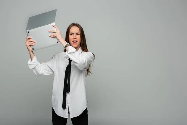 Angry young woman in white shirt with tie holding laptop isolated on grey — Stock Photo