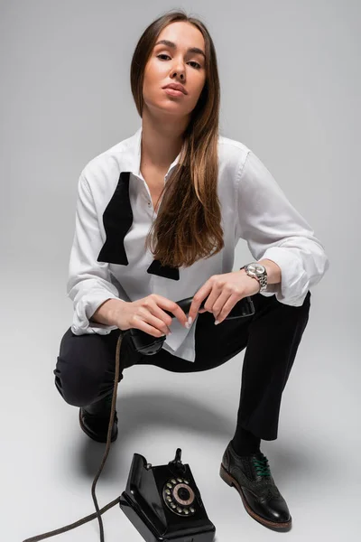 Brunette woman in white shirt with tie and pants holding retro telephone while sitting on grey — Stock Photo