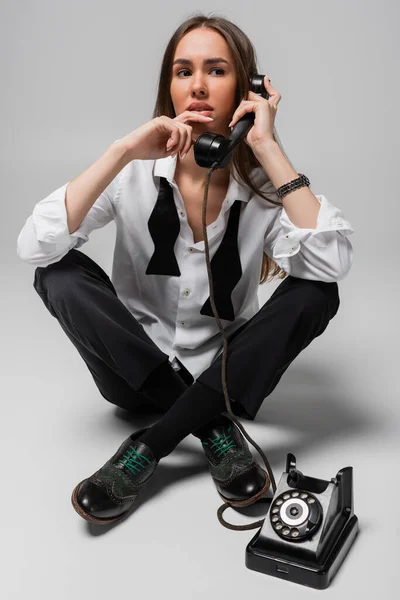 Pensive young woman in shirt with tie and pants using retro telephone while sitting on grey — Stock Photo