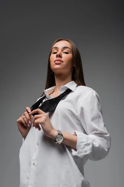 Portrait of confident woman in white shirt adjusting black tie while looking at camera isolated on grey, gender equality concept - foto de stock