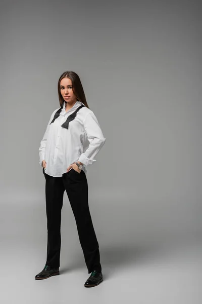 Full length of confident woman in shirt with tie and black pants standing with hands in pockets on grey, gender equality concept — Stockfoto