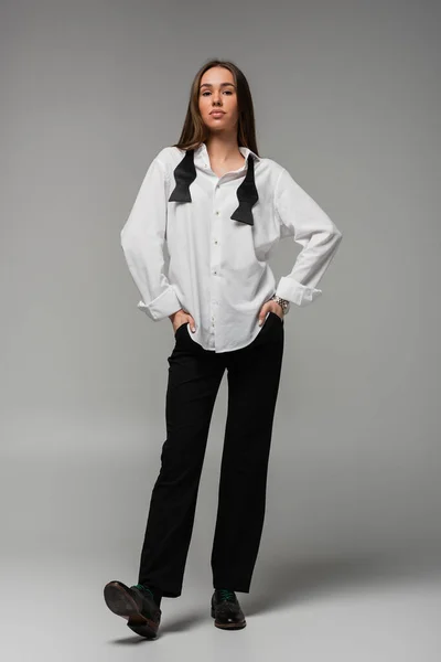 Повна довжина впевненої жінки в білій сорочці і штанях, що стоять руками в кишенях на сірому, концепції гендерної рівності — стокове фото