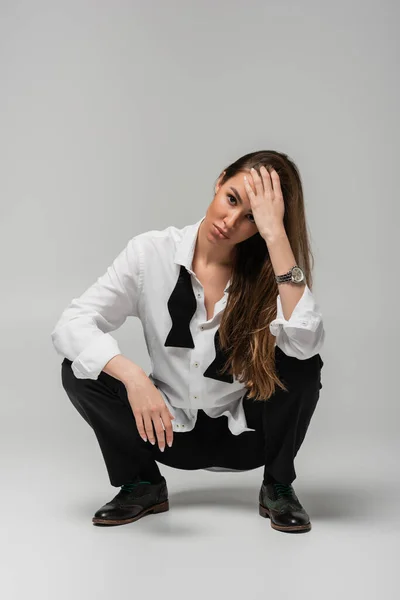 Full length of brunette woman in shirt with tie and black pants sitting on grey, gender equality concept — Foto stock
