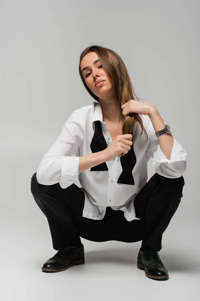 Pleine longueur de femme confiante en chemise avec cravate et pantalon noir ajustant les cheveux longs tout en étant assis sur gris, concept d'égalité des sexes — Photo de stock