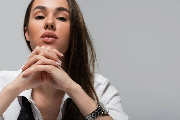 Portrait of brunette woman in white shirt looking at camera and sitting with clenched hands isolated on grey — Stock Photo