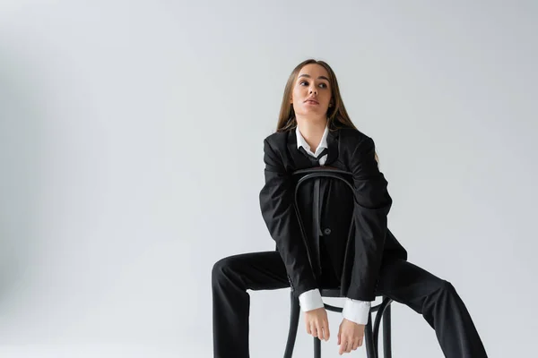 Young and brunette woman in suit sitting on wooden chair on grey background — Stock Photo