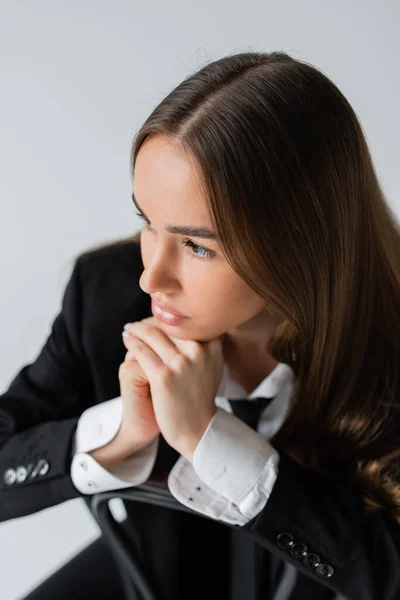 High angle view of young and brunette woman in suit leaning on wooden chair back isolated on grey — Stock Photo