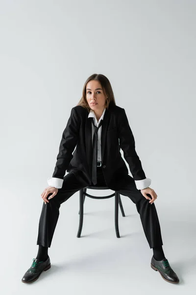 Full length of young brunette woman in black suit with tie sitting on wooden chair on grey — Stockfoto