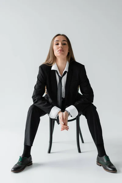 Full length of young confident woman in black suit with tie sitting on chair on grey — Stock Photo