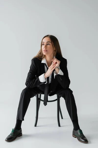 Full length of young brunette woman in black suit with tie sitting on wooden chair and looking away on grey — Stock Photo