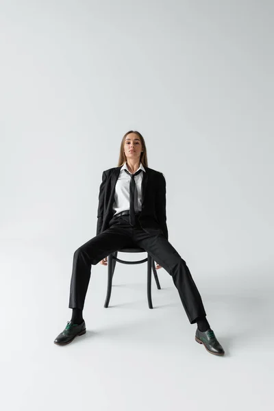 Full length of young brunette woman in black suit with tie sitting on wooden chair while looking at camera on grey — Stockfoto