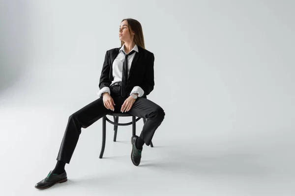 Full length of brunette woman in black suit with tie sitting on chair while looking away on grey — Stock Photo