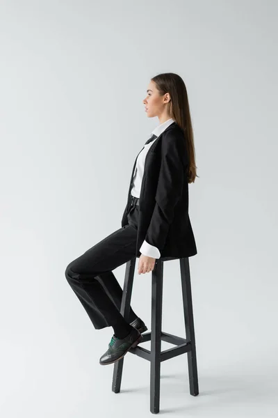 Side view of young brunette woman in black suit with tie sitting on wooden high chair on grey — Stock Photo