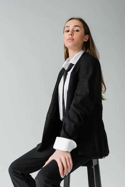Junge brünette Frau mit langen Haaren sitzt im schwarzen Anzug mit Krawatte und blickt vereinzelt in die Kamera auf grau — Stockfoto