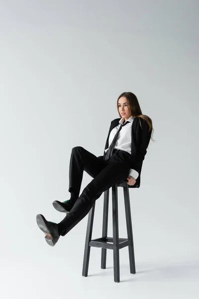 Full length of young brunette woman in black suit with tie sitting on high chair while posing on grey — Stock Photo