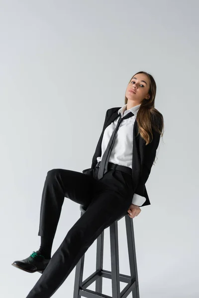 Full length of brunette woman in black suit with tie sitting on high chair and posing on grey — Stock Photo