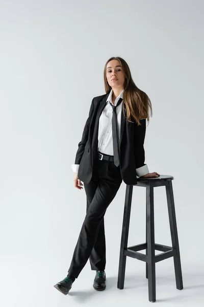 Full length of young brunette woman in black suit with tie standing near high chair on grey — Stock Photo
