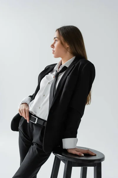 Side view of young brunette woman in black suit with tie standing near high chair on grey — Stock Photo