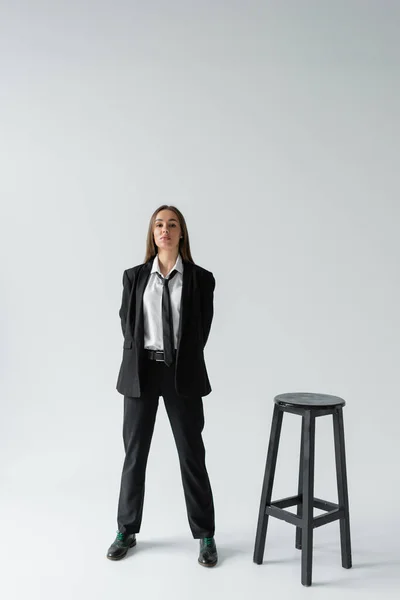 Повна довжина впевненої жінки в чорному костюмі з краваткою, що стоїть біля високого табурета на сірому — стокове фото