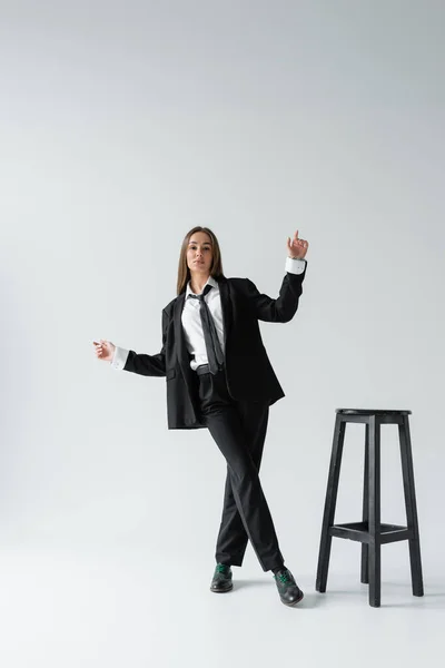 Full length of young brunette woman in black suit with tie posing near high chair on grey — Stock Photo