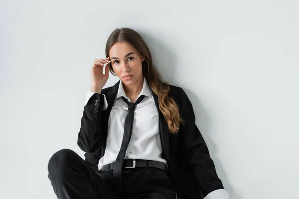 Brunette woman in black formal wear with tie looking at camera on grey background — Photo de stock