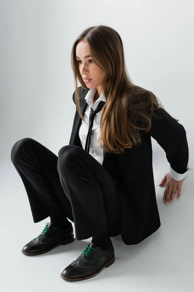 Full length of young and brunette woman in black formal wear with tie sitting on grey background — Fotografia de Stock
