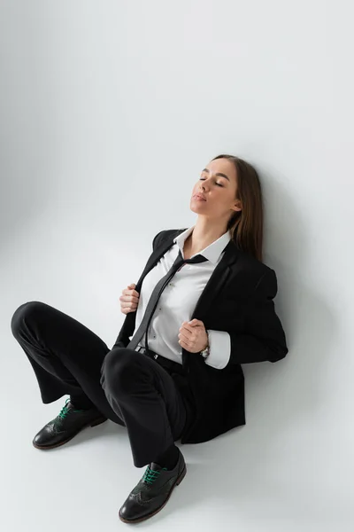 Full length of young and brunette woman in black formal wear sitting against white wall — Stock Photo