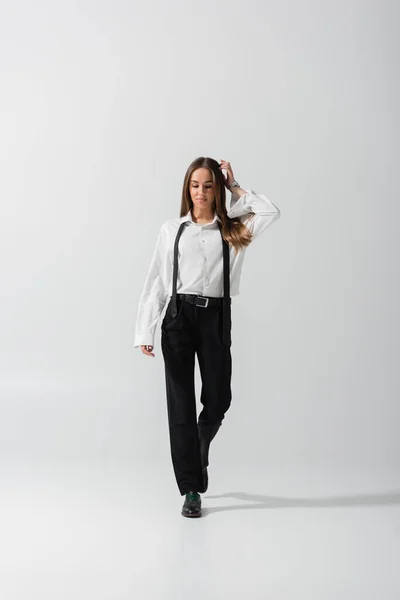 Full length of brunette woman in black pants and suspenders walking on grey — Stock Photo