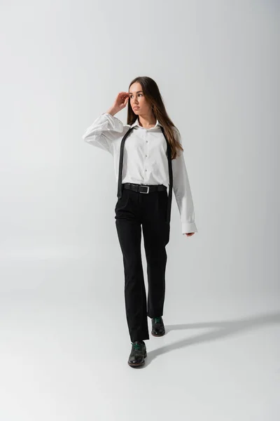 Full length of brunette young woman in black pants and suspenders walking on grey — Stock Photo