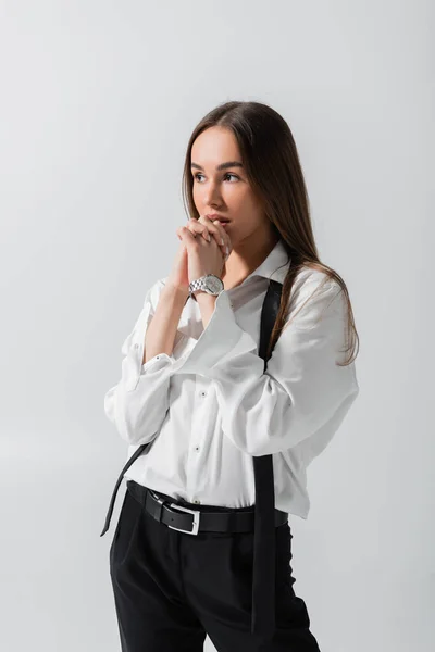 Pensive young woman in black pants and suspenders standing with clenched hands isolated on grey — Stock Photo