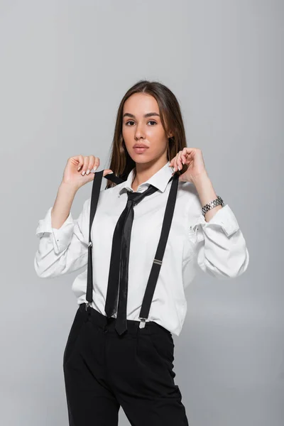 Brunette young woman in black pants and white shirt adjusting suspenders isolated on grey — Stock Photo
