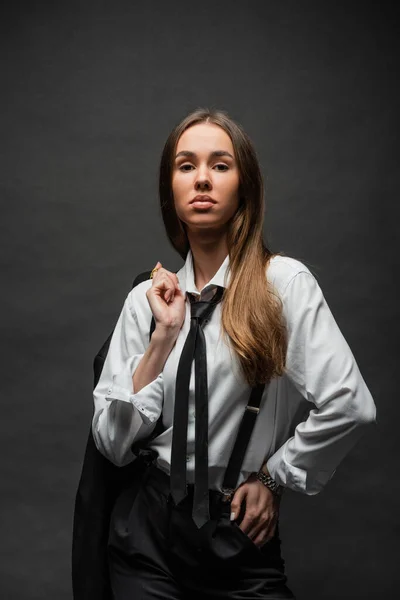 Morena mujer con el pelo largo de pie en el desgaste formal con tirantes mientras sostiene la chaqueta en negro - foto de stock