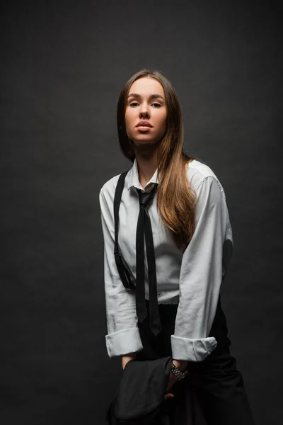 Brunette woman with long hair standing in suit with suspenders while holding blazer on black — Stock Photo
