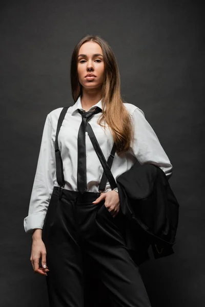 Brunette woman with long hair standing in suit with hand in pocket while holding blazer on black — Stock Photo