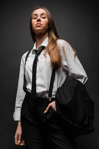 Confident woman with long hair standing in suit with hand in pocket while holding blazer on black — Stock Photo