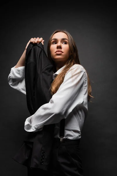 Low angle view of confident woman with long hair standing in suit and holding blazer on black — Stock Photo