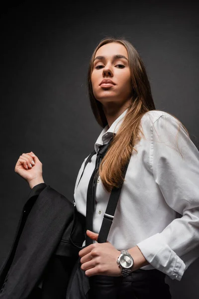 Low angle view of brunette woman in suit holding blazer and looking at camera on black — Stock Photo