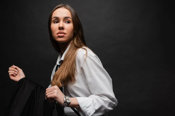 Young woman with brunette long hair standing in white shirt and holding blazer on black background — Stock Photo