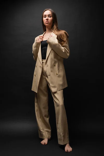 Full length of barefoot woman in beige formal wear standing and looking at camera on black — Stock Photo