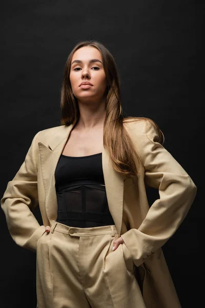 Brunette young woman in beige suit standing with hands in pockets on black — Stock Photo