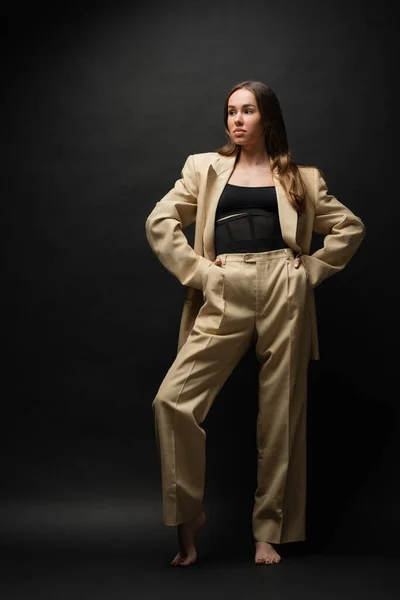 Full length of barefoot young woman in beige suit standing with hands in pockets on black — Foto stock