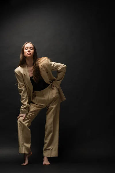 Full length of barefoot young woman in beige suit posing with hand on hip on black — Stock Photo