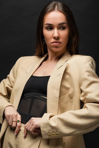 Brunette young woman in beige blazer adjusting trousers and looking away on black — Stock Photo