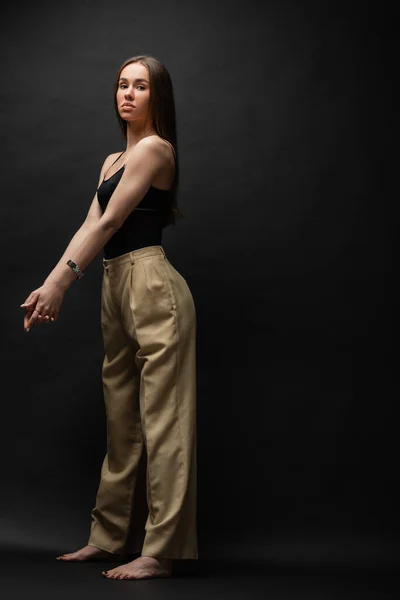 Full length of barefoot woman in top and beige pants standing with clenched hands on black background — Stock Photo