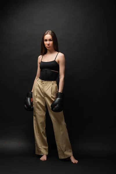 Full length of strong woman in top and beige pants posing in boxing gloves on black background — Stock Photo