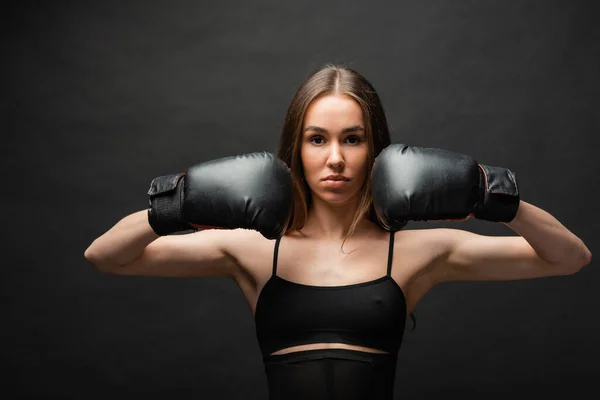 Brunette and strong young woman in top posing in boxing gloves on black background — Stock Photo
